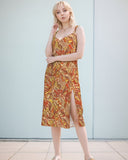  midi dress with a vibrant, psychedelic retro floral  print featuring swirling patterns in shades of red, yellow, and brown. The dress has a V-neckline, spaghetti straps, and a leg-baring side slit. Blissfully Brand